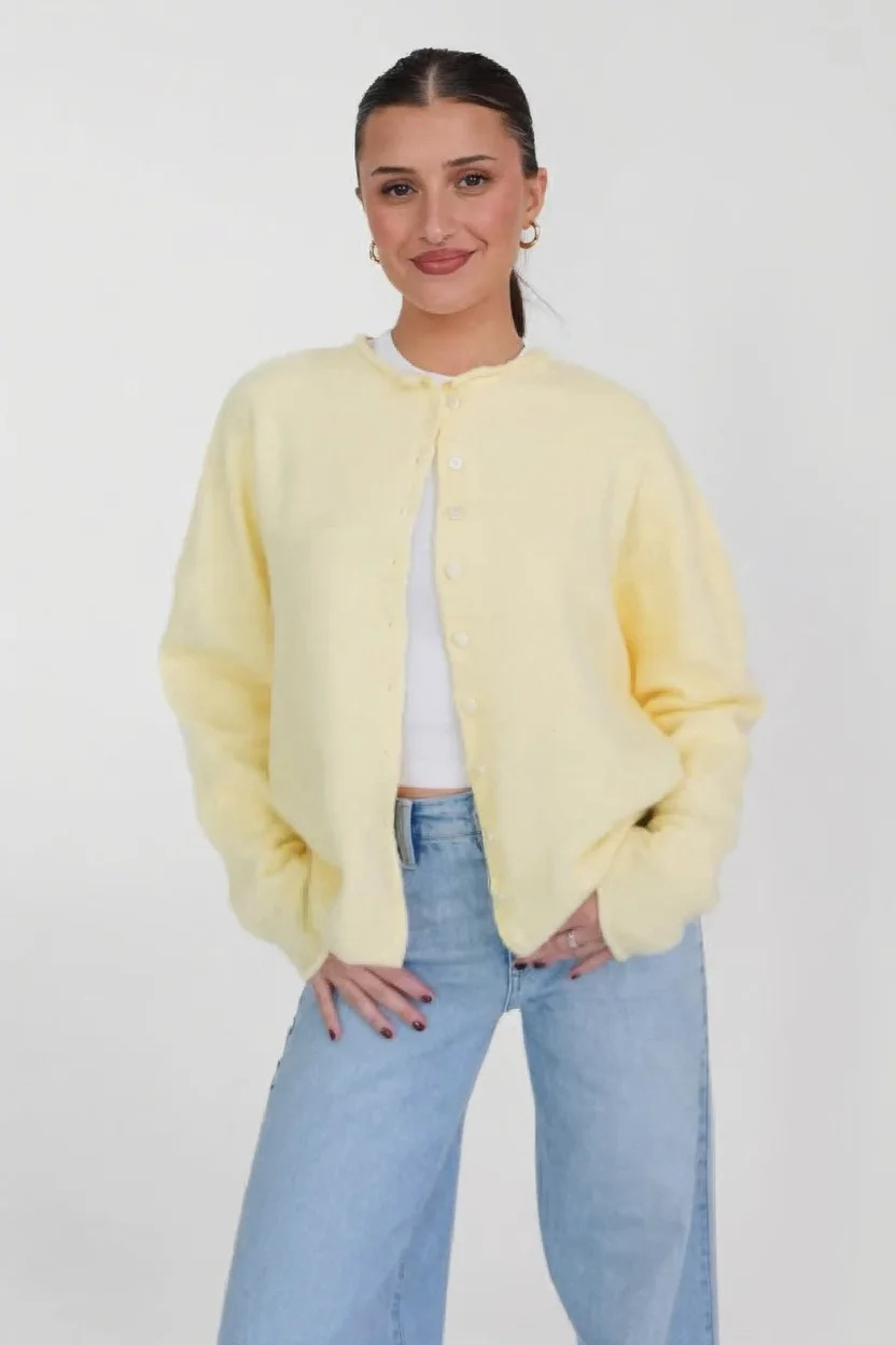 A woman with slicked-back hair in a pale yellow cardigan and wide-leg jeans, standing against a white studio wall. Body picture: pose, clothes and wall are kept.