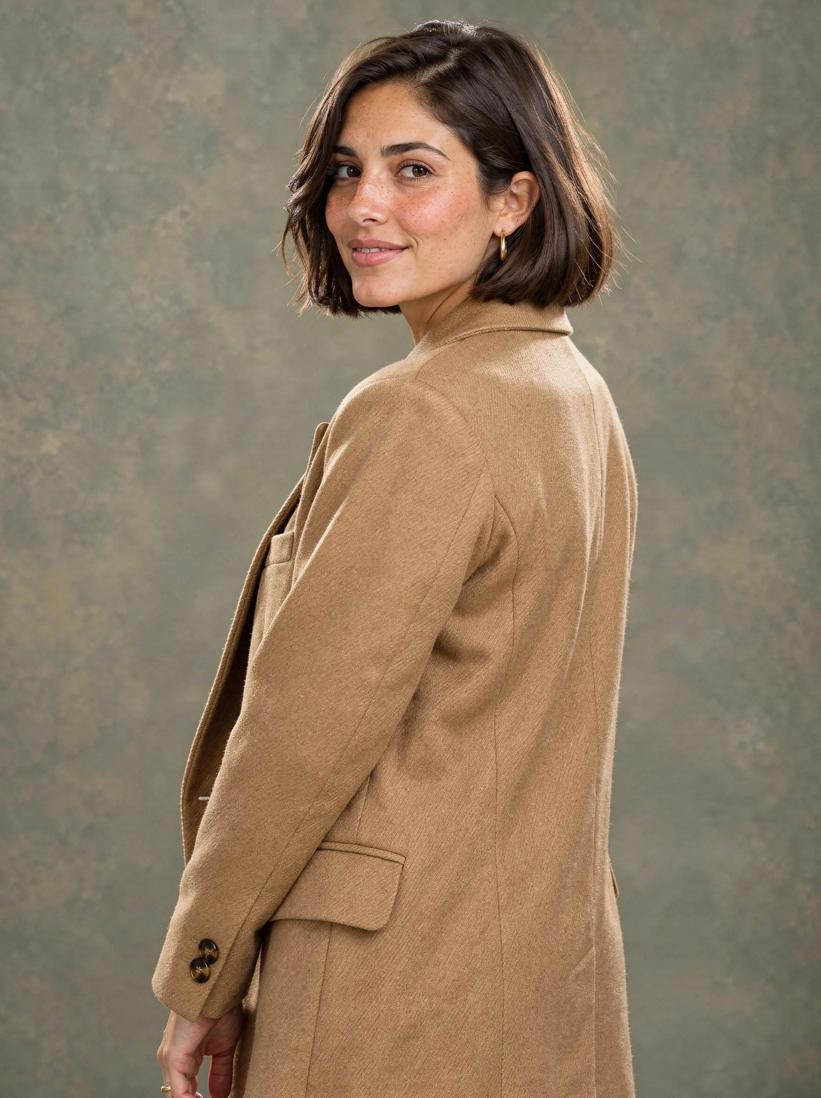 A woman with a chin-length dark brown bob and freckles, looking back over the shoulder of a camel wool coat, in front of a mottled olive studio backdrop