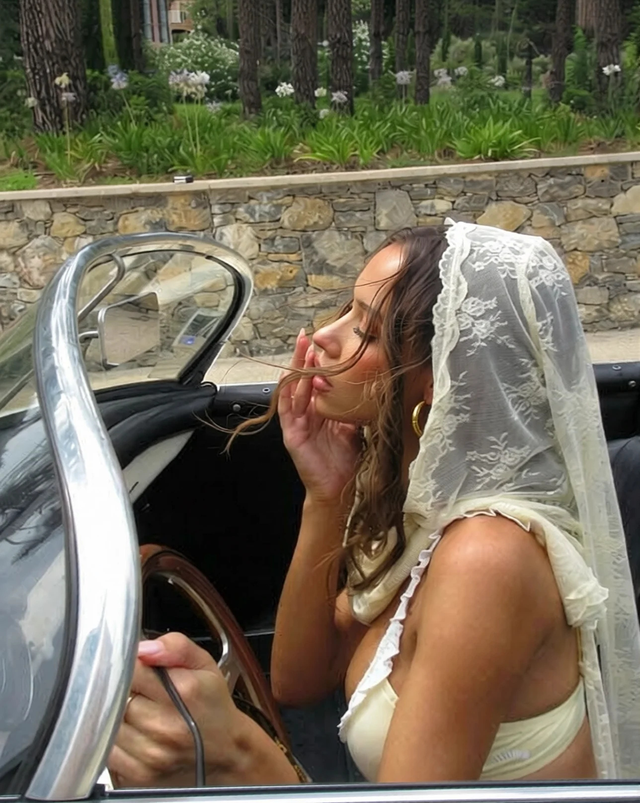 A woman in profile in an open-top classic car, a lace veil over her head, with a thick hank of windblown hair whipped across her forehead, nose and lips and loose wisps standing off her hairline