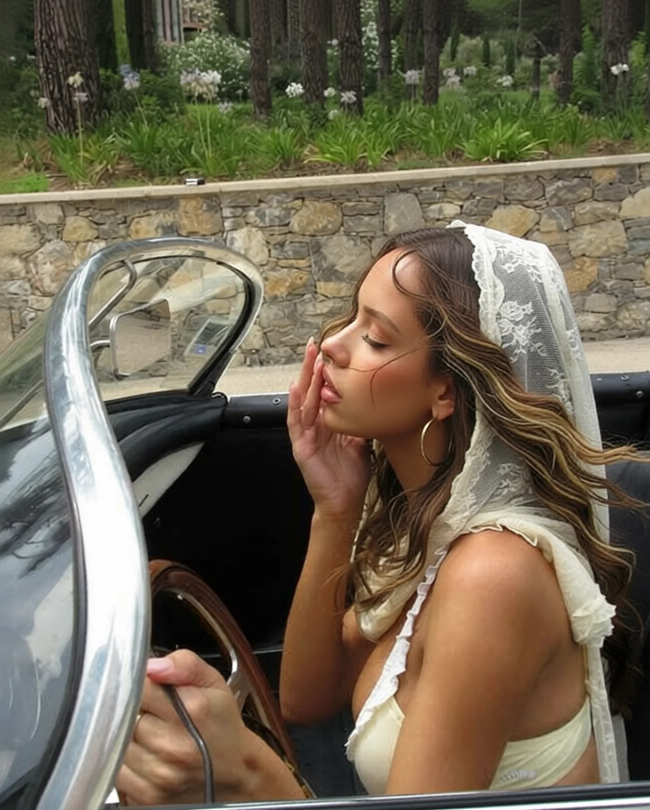 The same frame with the strands cleared off her face — the same woman, hand at her cheek, veil, car, stone wall and pine garden behind her