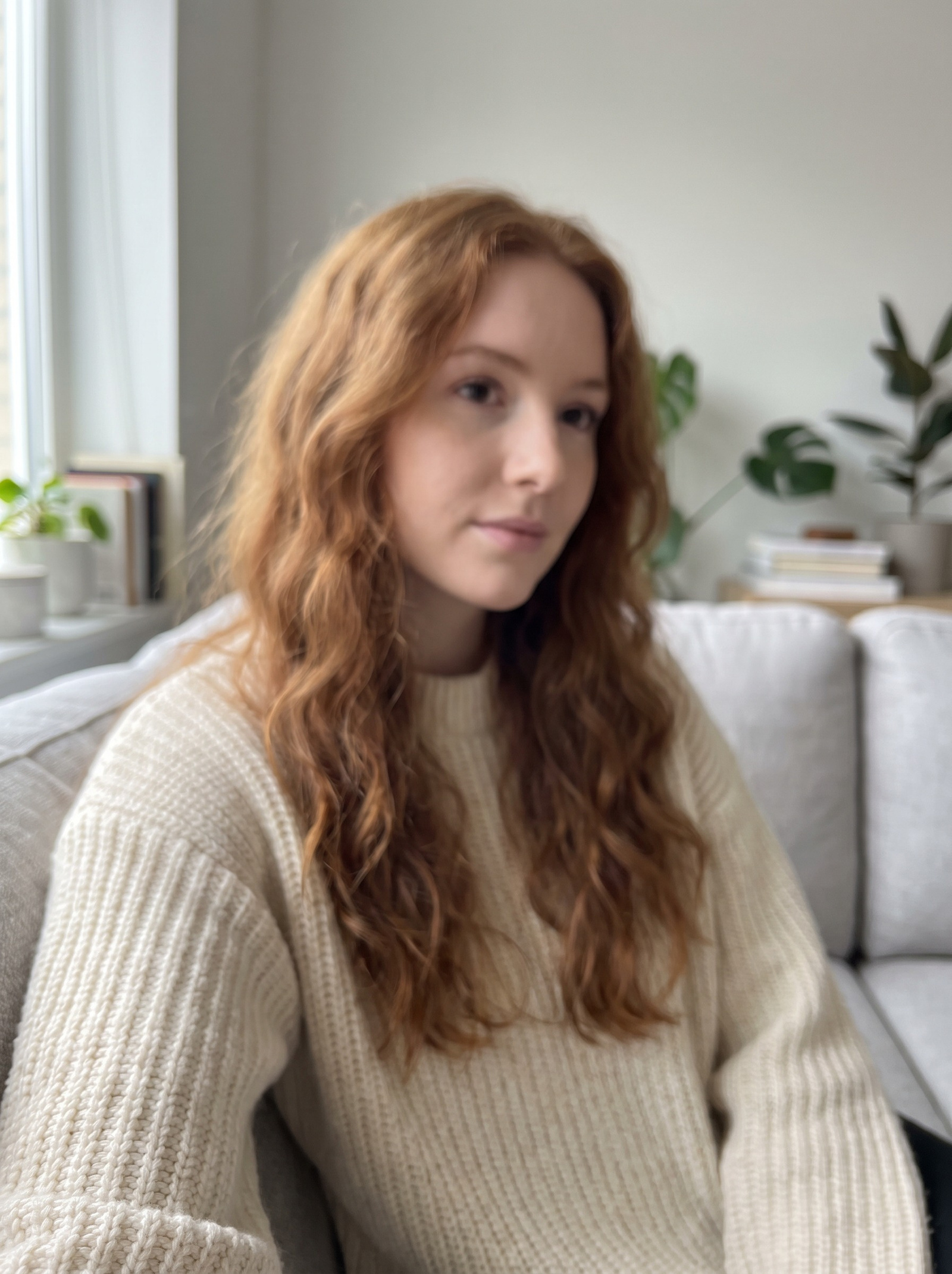 A soft, low-detail portrait: a woman in her twenties with long wavy auburn hair in a cream ribbed knit sweater, sitting on a pale sofa in window light — the skin smeared, the hair one undifferentiated mass, the eyes without crisp catchlights and the knit stitch unresolved