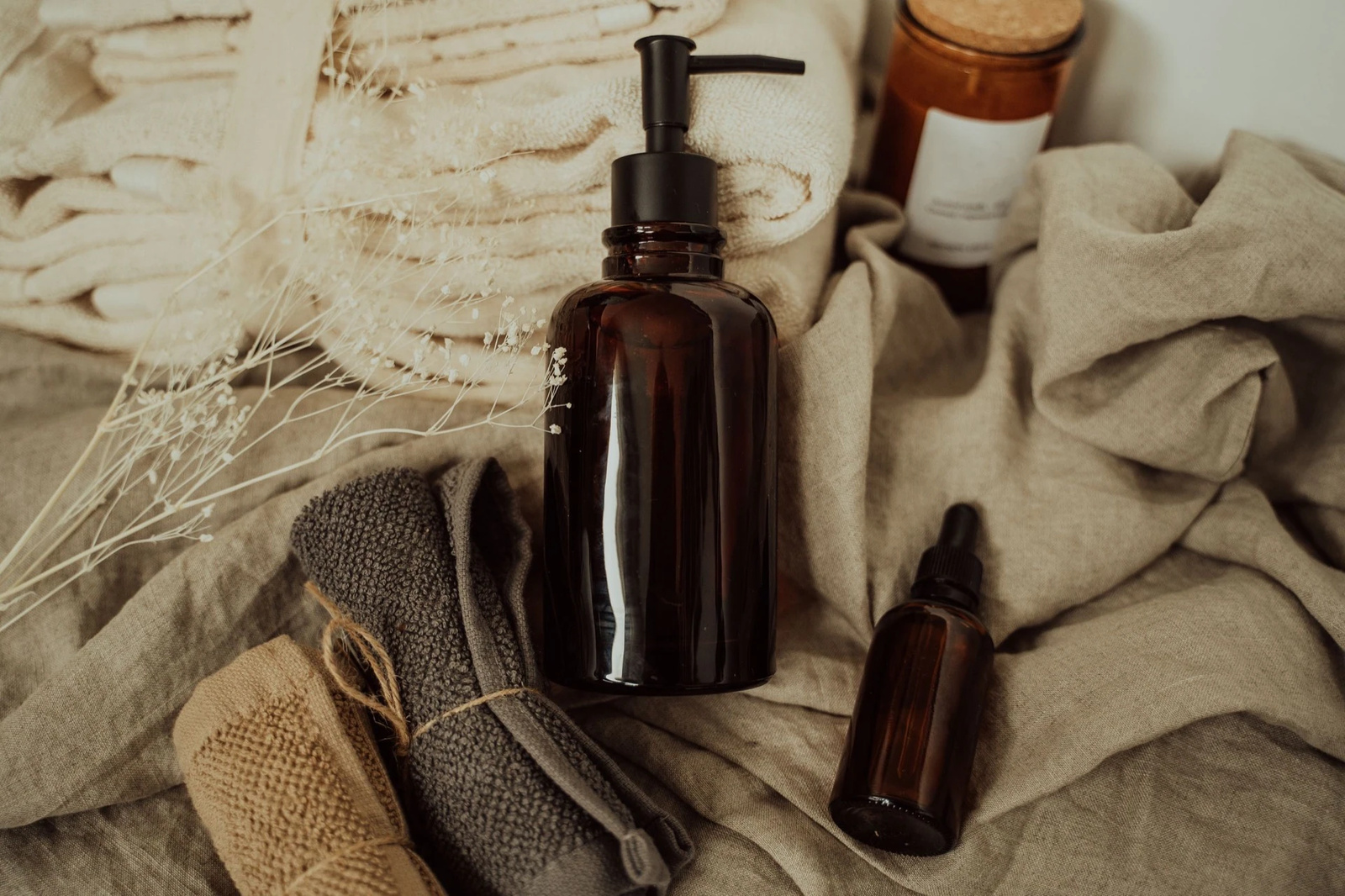 The product photo the reel is built from: an amber glass pump bottle with a black pump lying on rumpled linen beside a small amber dropper bottle, a cork-topped amber candle jar, folded cream towels, two rolled washcloths and dried gypsophila
