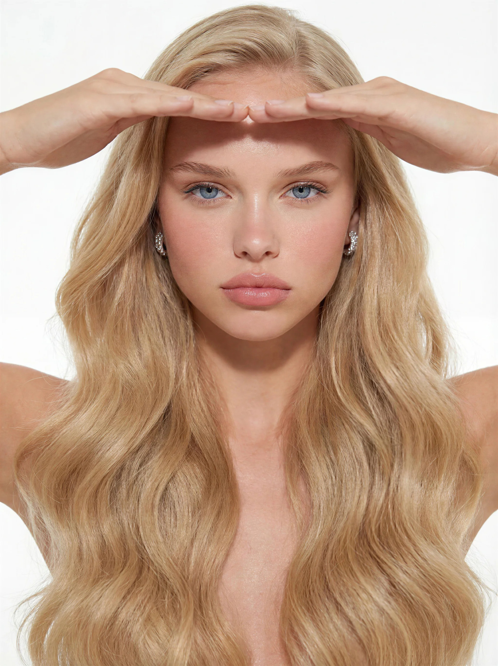 Studio beauty portrait of a blonde woman with long wavy hair, her hands framing her forehead against a bright white backdrop — the AI original, with poreless skin and a perfectly clean background