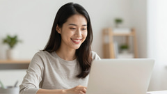Smiling woman with long dark hair in a grey sweater typing on a laptop in a bright room with plants