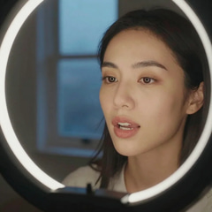 Close-up of a woman's face lit by soft ring light in a home studio, lips parted mid-syllable as she speaks to an off-frame camera, warm skin