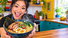 Excited cook holding up a steaming bowl of ramen in a bright kitchen