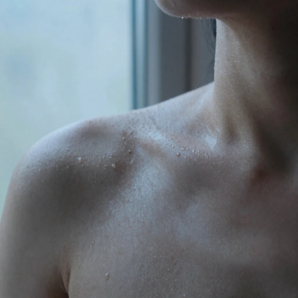Macro of dewy skin across a bare shoulder and collarbone, tiny water droplets and a few small moles in cool blue window light