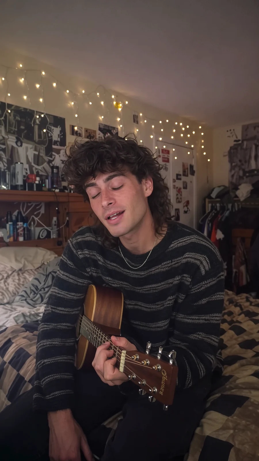 A young man with a mullet sings with an acoustic guitar on his unmade bed under fairy lights, camcorder look