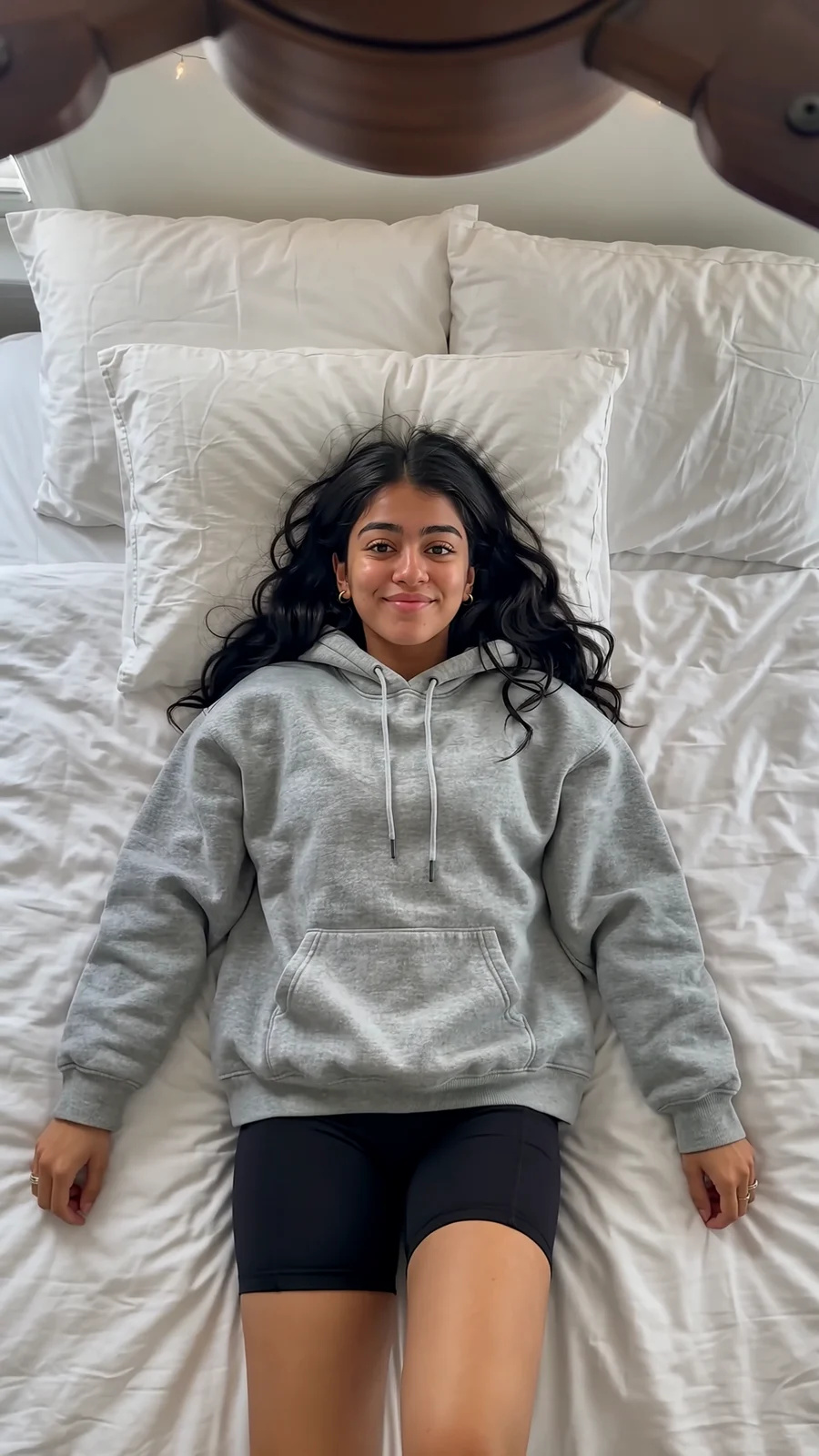 Overhead shot of a young woman lying on her bed; a wooden ceiling-fan blade sweeps across the lens and each pass changes her outfit, from a grey hoodie to a leather bomber with cargo jeans to a brown knit mini dress.