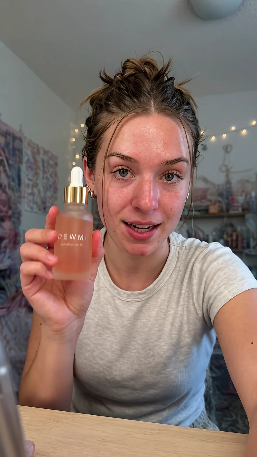 A young woman at her desk shows a DEWMI serum bottle to the camera and pats it on her cheek