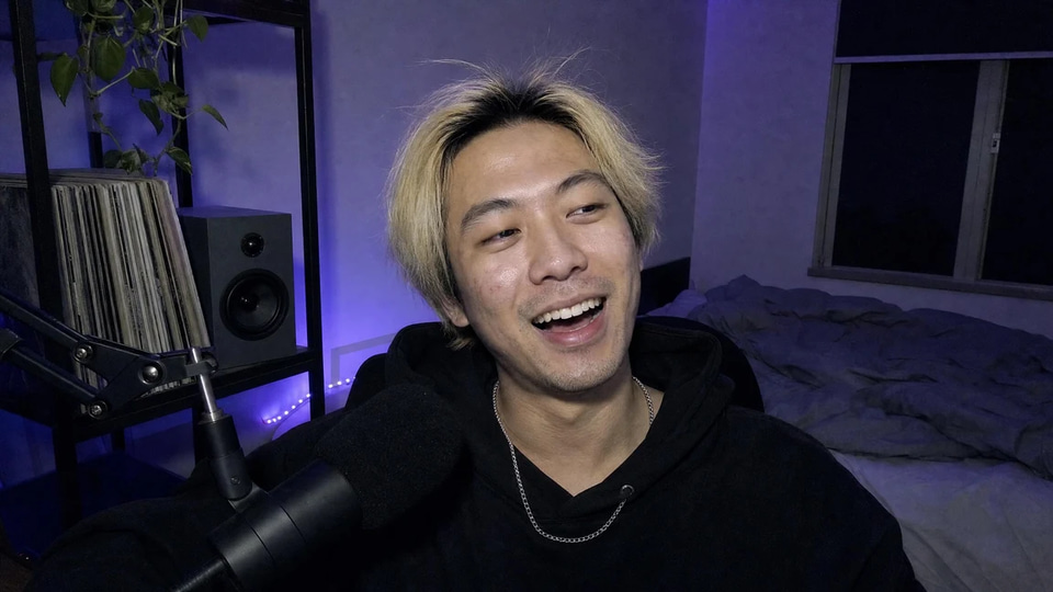 Webcam frame of a young man with bleached hair laughing on a late-night stream, violet LED light on the wall behind and his face lit evenly