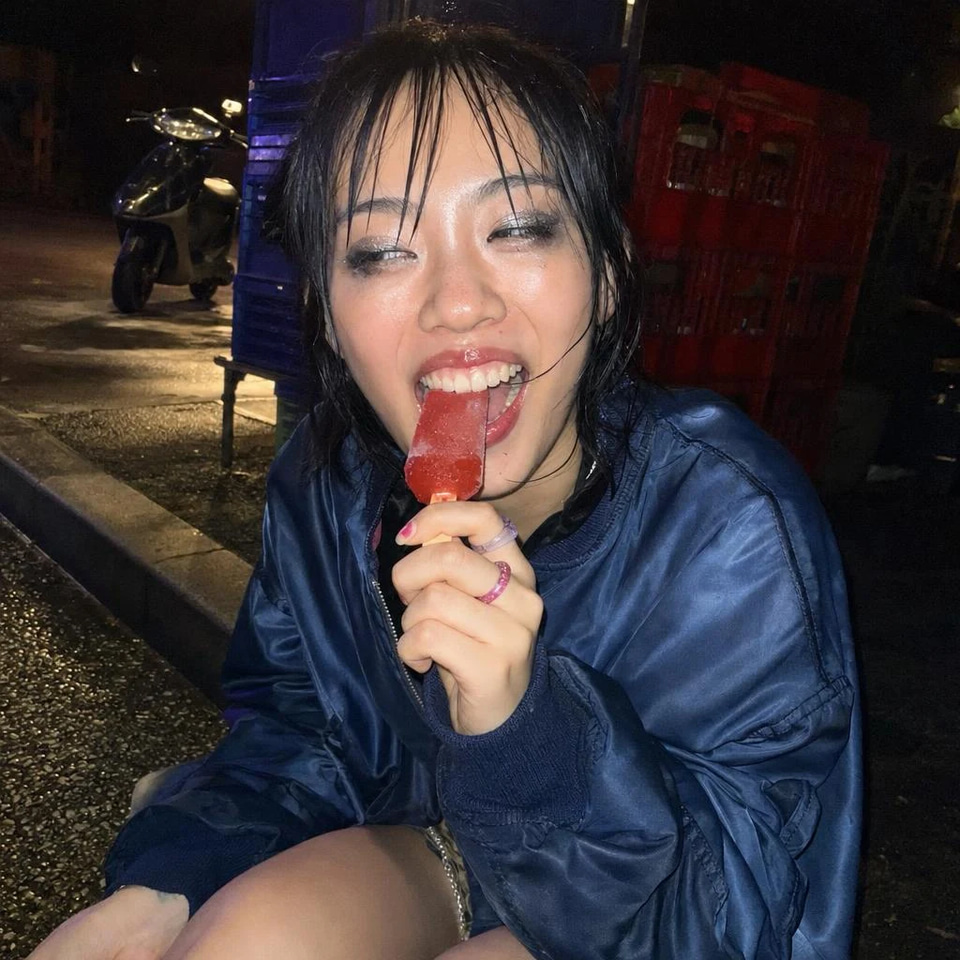Rainy Letters, a late-night pop playlist in English: A young woman with a wet black bob and glitter eyeliner laughs while biting a red ice lolly on a wet kerb at night, lit by phone flash.