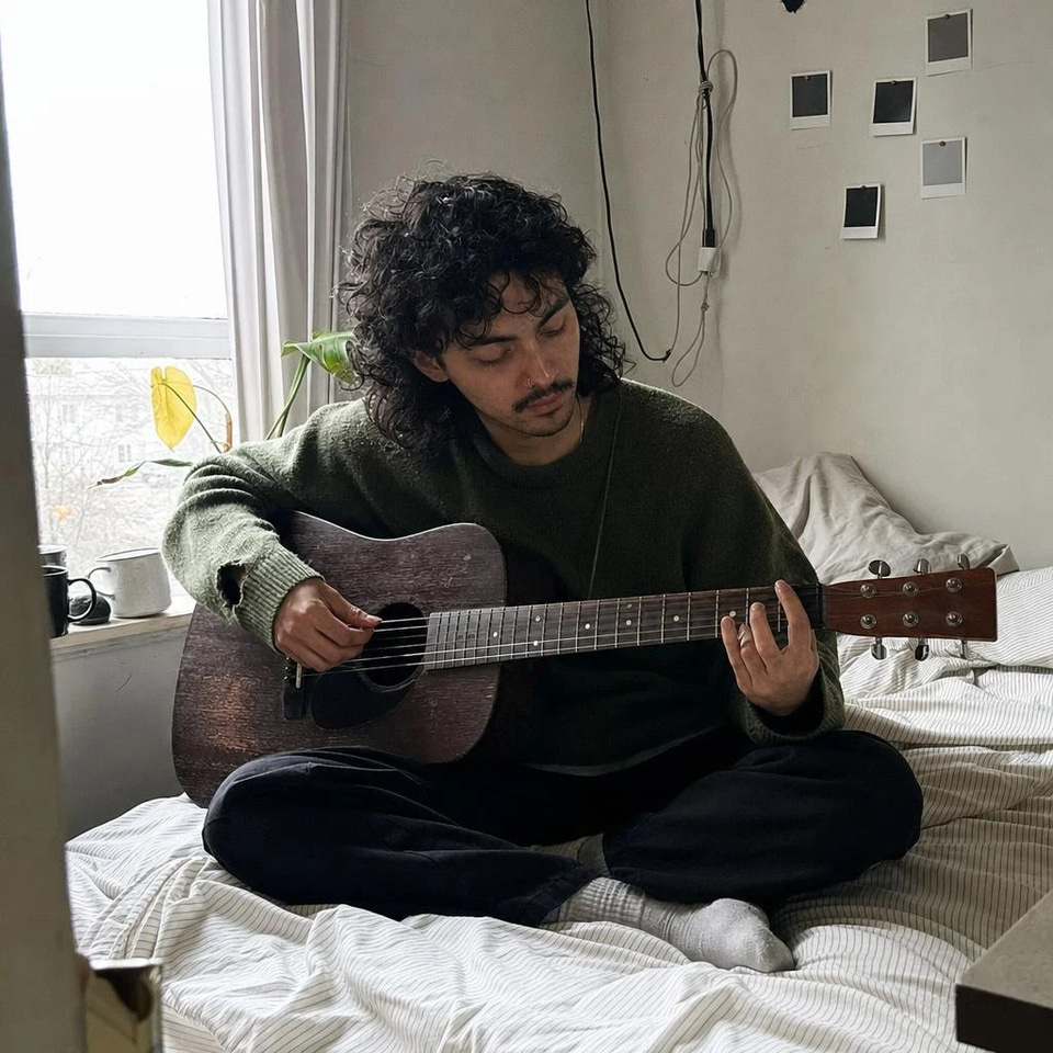 Quiet Rain Library, a lo-fi study playlist in English: A young man with a curly mullet sits cross-legged on an unmade bed playing a worn acoustic guitar in grey window light, with blank polaroids on the wall.