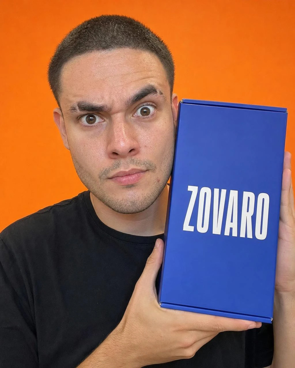 A 24-year-old man with a buzz cut, mouth closed and one eyebrow raised, holding a cobalt ZOVARO box beside his face against an orange wall