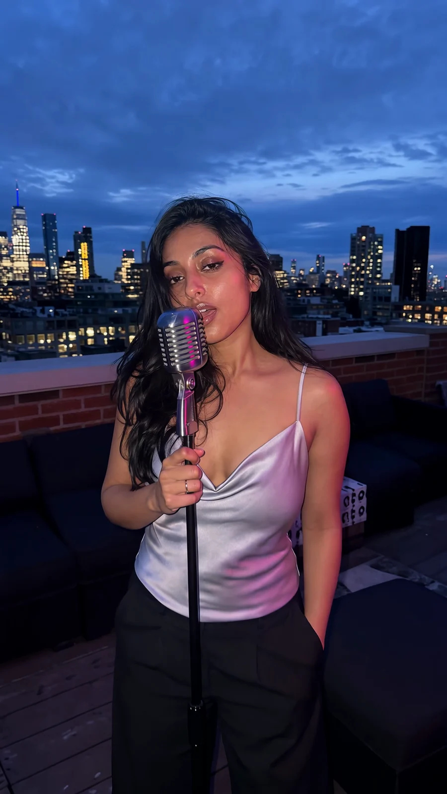 A young woman in a silver camisole sings into a vintage microphone on a blue-hour rooftop as the camera pushes in
