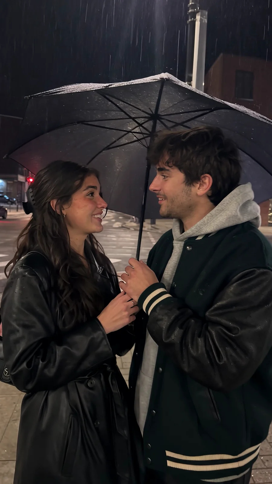 A young adult couple in coats share an umbrella on a rainy street corner; the wind flips it, she drops it and they kiss