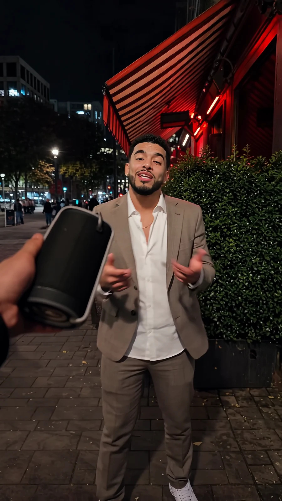 A young man in a tan checked suit freestyles beside a portable speaker under red neon outside a restaurant while passers-by film