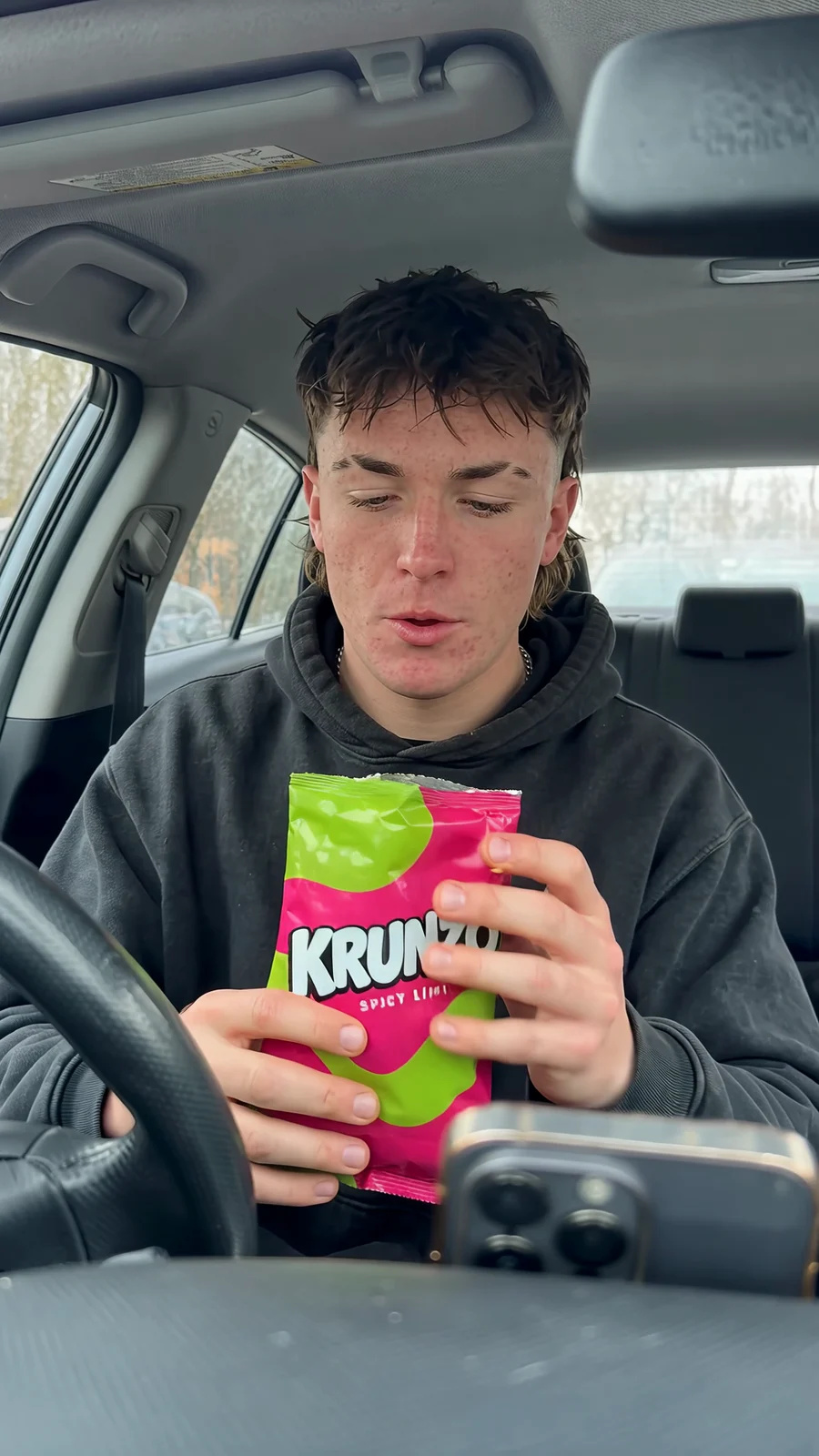 A young man in a car opens a pink-and-green KRUNZO snack bag, takes a bite and reacts to the dashboard camera