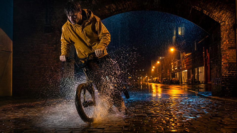 A cyclist in a mustard raincoat skids to a stop under a brick arch in heavy rain at night