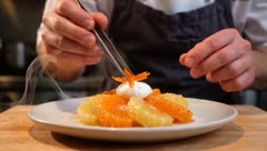 Chef plating a citrus dessert with tweezers in a restaurant kitchen
